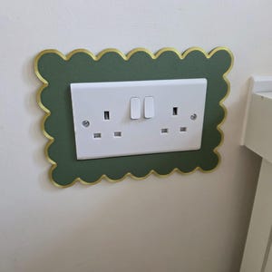 Pode incluir: Uma placa de tomada elétrica dupla branca emoldurada por uma cobertura retangular verde-escura com borda recortada e acabamento dourado. A tomada possui dois interruptores e dois conjuntos de três orifícios, fixada por dois parafusos.