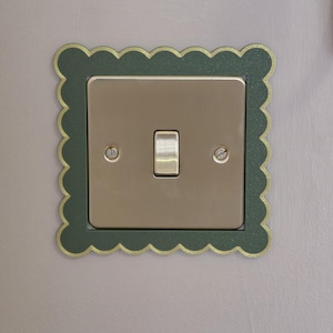 Puede incluir: Una placa de interruptor de luz metálica con un interruptor de palanca central y dos tornillos de cabeza plana, enmarcada por un borde festoneado de color verde oscuro con un borde dorado. La placa cuadrada tiene un acabado liso y brillante.