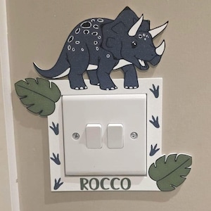 May include: A light switch surround with a dark blue triceratops dinosaur design. The white surround has green leaf and footprint details. The name "ROCCO" is printed in green at the bottom, adding a personalised touch.