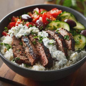 Może przedstawiać: Ciemnoszara miska wypełniona kolorową sałatką i pokrojoną stekiem. Sałatka zawiera pomidory, oliwki i og&oacute;rki. Stek jest pokrojony i polany kremowym sosem i ziołami, podawany na białym ryżu.