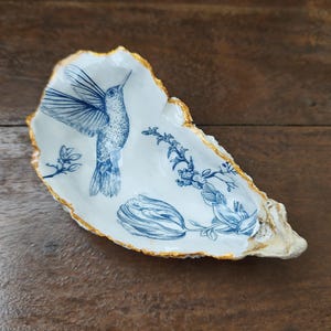 Könnte beinhalten: Dekorative Austernschalen-Schale mit blauem Kolibri- und Blumenmuster. Das weiße Innere hat blaue Illustrationen und einen goldfarbenen Rand. Die Schale liegt auf einer dunklen Holzoberfläche.