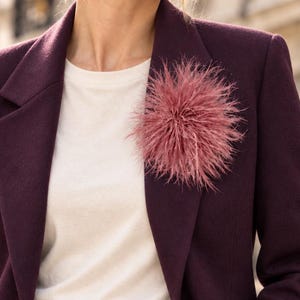 Könnte beinhalten: Ein tiefvioletter Blazer über einem schlichten weißen T-Shirt mit einer großen, runden, altrosa Federbrosche am linken Revers. Der Blazer hat einen fallenden Reverskragen, strukturierte Schultern und einen einzelnen Knopfverschluss.