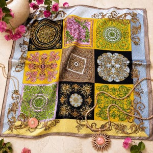 Könnte beinhalten: Ein quadratisches Seidentuch mit einem mehrfarbigen Gittermuster und verzierten barocken Motiven in Gold, Schwarz, Grün und Pink. Zwei goldfarbene Halsketten mit runden, pinkfarbenen Anhängern in Sternform liegen auf dem Stoff.