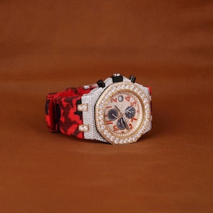 May include: A luxury wristwatch with a red and black camouflage patterned strap. The watch face is gold with black sub-dials and a diamond-encrusted bezel. A striking timepiece.