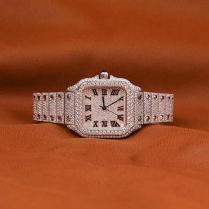 May include: A silver wristwatch with a square face and a matching band. The watch face features black Roman numerals and black hands. The watch and band are encrusted with small, sparkling stones. The watch is displayed on a brown surface.