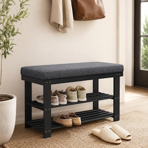 May include: A black wooden shoe bench featuring a rectangular dark gray upholstered seat cushion. The bench has two slatted shelves for storage, with four pairs of shoes placed on the shelves and one pair of beige slides on the floor.