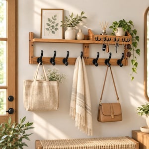 May include: A wall-mounted wooden shelf featuring a top ledge for decor and a lower section with eight black metal hooks. The shelf holds a framed botanical print, two ceramic vases, a reed diffuser, and a trailing green plant.