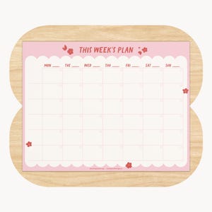 May include: A pink weekly planner with the text "THIS WEEK'S PLAN" and a grid for daily scheduling. The planner has a scalloped edge and is decorated with small red flower illustrations. It is set on a wooden background.