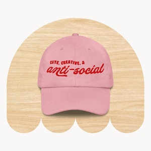 May include: A light pink baseball cap featuring the embroidered red text "CUTE, CREATIVE, & anti-social" across the front panel. The cap has a structured crown, a curved brim, and a button at the top center, showcasing a clean and simple design.