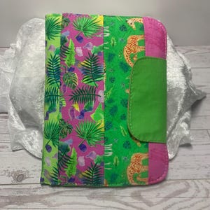Puede incluir: Una funda de diario de tela con un estampado tropical vibrante. La funda presenta un diseño de patchwork con paneles rosas, verdes y amarillos. Un panel tiene un estampado de leopardo. Una solapa verde asegura el diario.