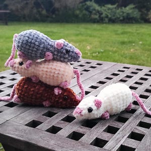 May include: Four small crocheted plush mice made from soft yarn. Three mice are stacked vertically in gray, light pink, and dark brown, while one white mouse sits to the right. Each mouse features small pink ears, pink feet, and black bead eyes.