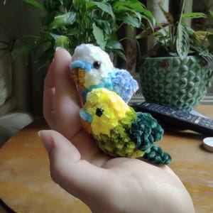 Puede incluir: Dos pequeños pájaros de peluche hechos a mano con hilo suave y difuso. Un pájaro tiene la cabeza blanca con alas azules y azul claro, mientras que el otro tiene la cabeza amarilla con alas verdes y verde oscuro. Ambos tienen ojos de cuentas negras.