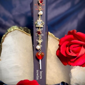 May include: A gold-colored bookmark with red heart and crown charms, pearls, and a lollipop charm. The bookmark is placed in a book with the title "had been wish" by Laura Nowlin. A red rose is in the background.