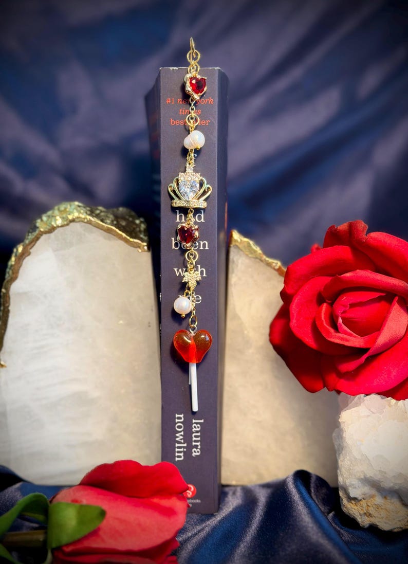 May include: A gold-colored bookmark with red heart and crown charms, pearls, and a lollipop charm. The bookmark is placed in a book with the title "had been wish" by Laura Nowlin. A red rose is in the background.