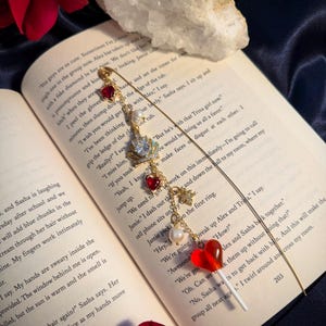 May include: A gold-toned bookmark with dangling charms, including red heart-shaped beads, a pearl, and a crown-like embellishment. The bookmark rests on an open book with visible text and a red rose.