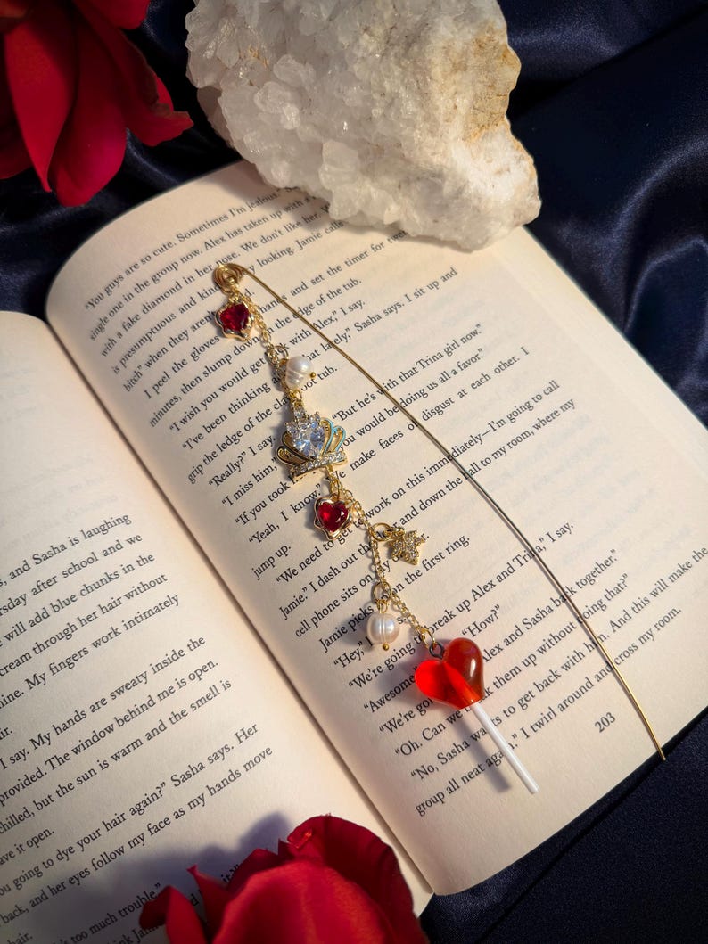 May include: A gold-toned bookmark with dangling charms, including red heart-shaped beads, a pearl, and a crown-like embellishment. The bookmark rests on an open book with visible text and a red rose.
