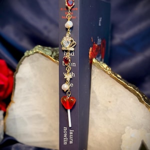 May include: A gold-colored bookmark with red heart-shaped gems, pearls, and a crown charm. The bookmark is attached to a dark blue book with the title "had been with" and the author's name, Laura Nowlin.