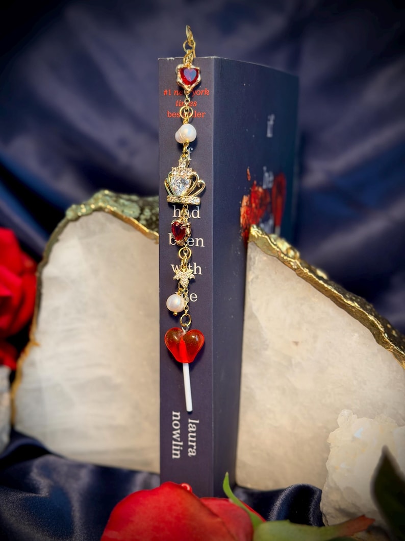 May include: A gold-colored bookmark with red heart-shaped gems, pearls, and a crown charm. The bookmark is attached to a dark blue book with the title "had been with" and the author's name, Laura Nowlin.