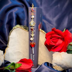 May include: A decorative bookmark with a gold chain and charms, including a red heart, crown, and pearls, is placed in a book. A red rose and white stones are in the foreground. The book's title is partially visible.