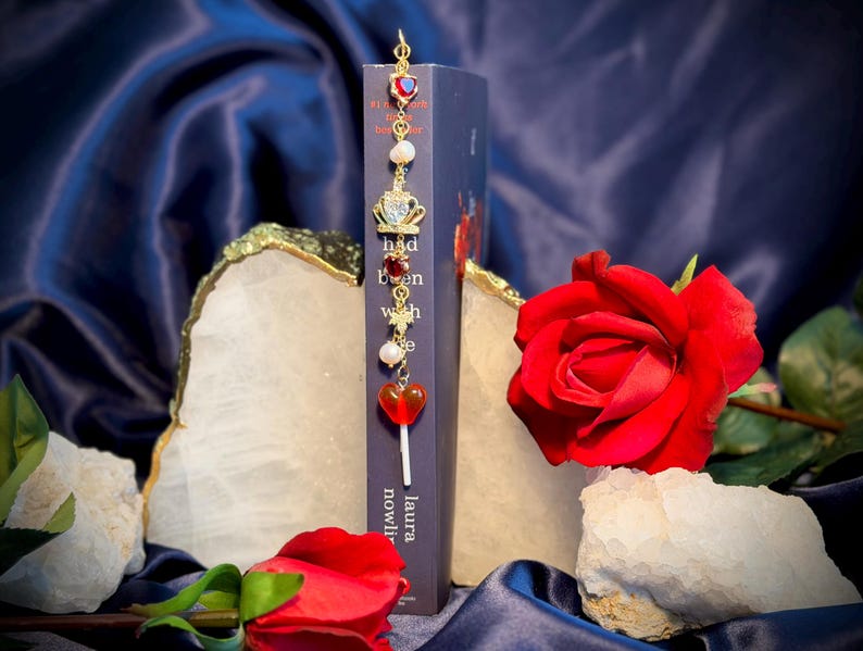 May include: A decorative bookmark with a gold chain and charms, including a red heart, crown, and pearls, is placed in a book. A red rose and white stones are in the foreground. The book's title is partially visible.