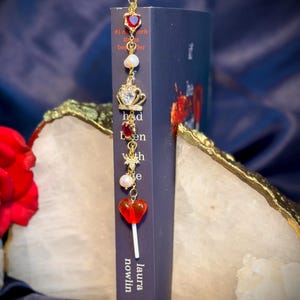 May include: A decorative bookmark with a gold chain and charms, including a red heart, a crown, and a lollipop. The bookmark is placed in a book with the title "Had Been With Me" by Laura Nowlin.
