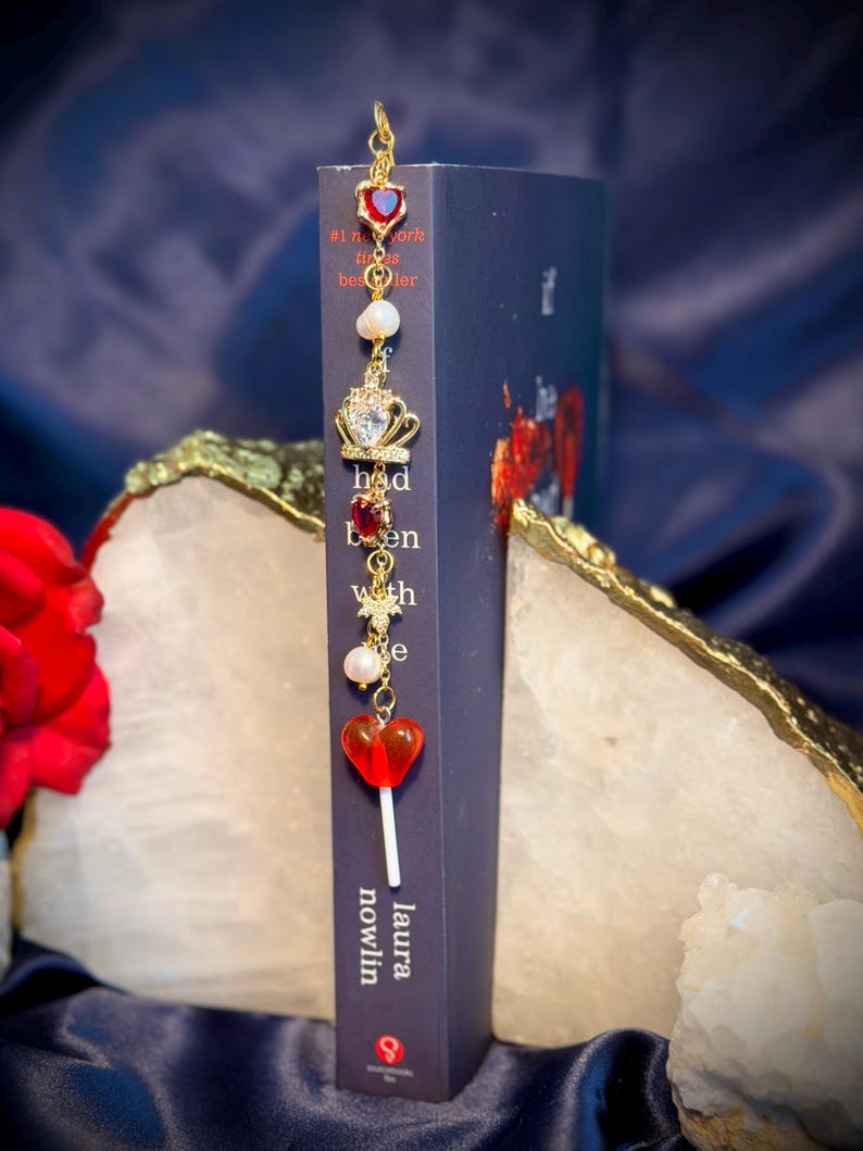 May include: A decorative bookmark with a gold chain and charms, including a red heart, a crown, and a lollipop. The bookmark is placed in a book with the title "Had Been With Me" by Laura Nowlin.