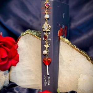 May include: A gold-colored bookmark with a series of charms, including a heart-shaped gem, a pearl, and a crown, hangs from the top of a book. The book is dark blue with the title "had been with" and the author's name, "laura nowlin". A red heart-shaped lollipop charm is also attached.
