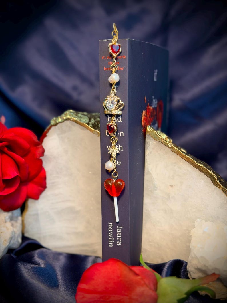May include: A gold-colored bookmark with a series of charms, including a heart-shaped gem, a pearl, and a crown, hangs from the top of a book. The book is dark blue with the title "had been with" and the author's name, "laura nowlin". A red heart-shaped lollipop charm is also attached.