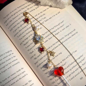 May include: A gold-toned bookmark with charms, including red hearts, a crown, pearls, and a lollipop. The bookmark is placed on an open book with printed text. The overall aesthetic is romantic and whimsical.