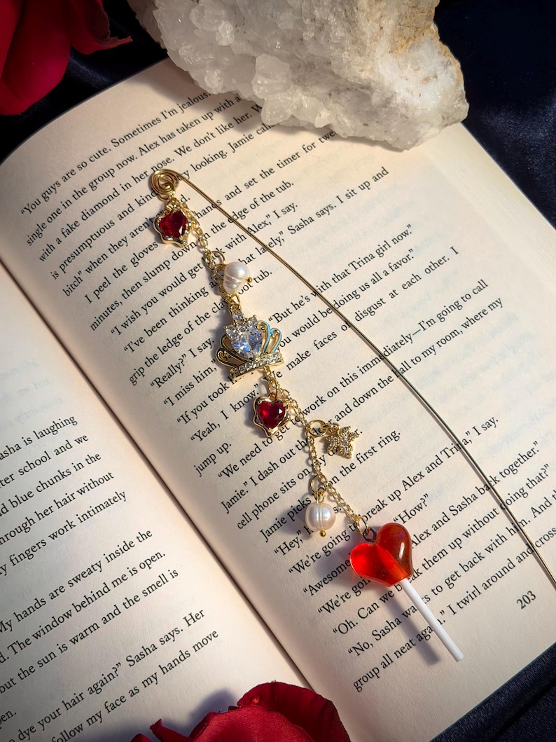 May include: A gold-toned bookmark with charms, including red hearts, a crown, pearls, and a lollipop. The bookmark is placed on an open book with printed text. The overall aesthetic is romantic and whimsical.