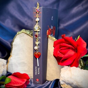 May include: A decorative bookmark with red heart and crown charms, pearls, and a gold-colored hook. The bookmark is placed in a dark blue book with the author's name, Laura Nowlin, visible. Red roses and quartz crystals are in the background.