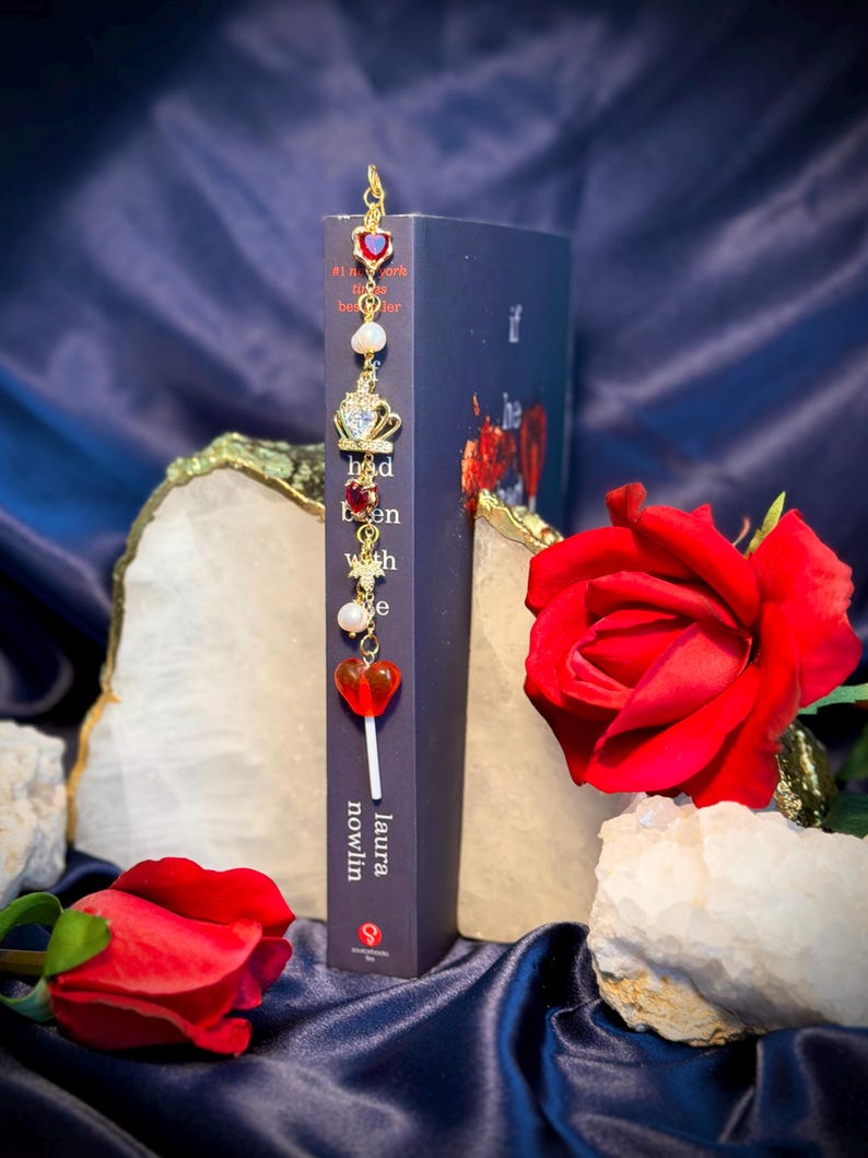 May include: A decorative bookmark with red heart and crown charms, pearls, and a gold-colored hook. The bookmark is placed in a dark blue book with the author's name, Laura Nowlin, visible. Red roses and quartz crystals are in the background.