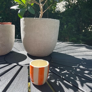 Könnte beinhalten: Ein kleiner Keramikbecher mit vertikalen orangefarbenen und weißen Streifen. Der Becher hat eine glatte, glänzende Oberfläche und eine zylindrische Form. Er steht vor zwei großen, strukturierten, hellgrauen Betonpflanzgefäßen mit grünen Pflanzen.