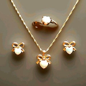 May include: A gold-coloured jewellery set comprising a necklace, earrings, and a ring. The necklace features a heart-shaped pendant with a bow design. The earrings and ring also have heart-shaped stones. The jewellery is displayed on a grey background.