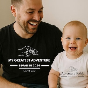 May include: A smiling adult and baby wearing matching black and white t-shirts. The black shirt reads "MY GREATEST ADVENTURE BEGAN IN 2026 LIAM'S DAD" with a fist bump graphic. The baby's shirt says "Adventure Buddy Since 2026" with a similar graphic.