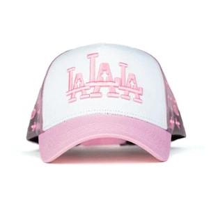 Peut inclure: Casquette de baseball blanche et rose avec le texte "LA LA LA" brod&eacute; en rose sur le devant. La casquette a une visi&egrave;re rose et un dos en filet avec un motif de croix roses.