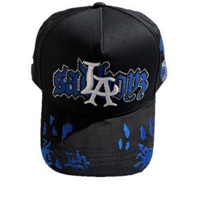 Puede incluir: Gorra de b&eacute;isbol negra con el texto "LA" en bordado blanco y azul. La visera presenta detalles decorativos azules. La gorra tiene una visera curva y un bot&oacute;n en la parte superior.