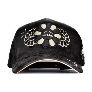 May include: Black baseball cap with a velvet texture. The cap features a floral design with a sad face in the center and two smaller flowers on the sides. The flowers are black and cream-colored, with white accents.