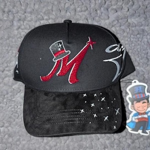 May include: Black baseball cap with a suede bill. The cap features a red and white embroidered "M" with a top hat design. White stars are embroidered on the bill. A tag with a cartoon magician is attached.