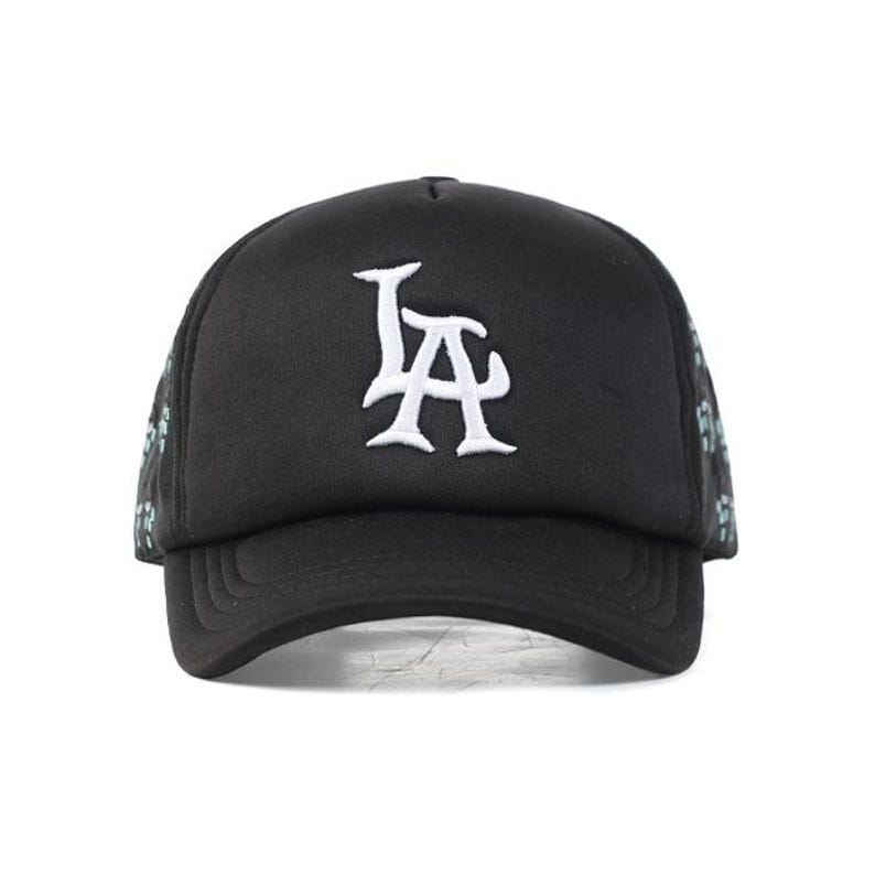 Peut inclure: Casquette de camionneur noire avec le logo "LA" blanc brod&eacute; sur le devant. La casquette a une visi&egrave;re incurv&eacute;e et un dos en filet avec un motif floral bleu et vert.