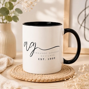 Puede incluir: Una taza de cerámica blanca con interior y asa negros. La taza presenta el texto "AG" en escritura cursiva, junto con "Armando Gonzales Designer EST. 1999" en negro. La taza está sobre un posavasos redondo y tejido.
