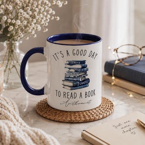 Puede incluir: Una taza de cerámica blanca con interior y asa azules, con la frase "IT'S A GOOD DAY TO READ A BOOK" y una pila de libros azules. La taza está sobre un posavasos tejido.