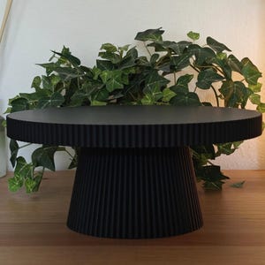 Puede incluir: Un soporte para pasteles negro con borde acanalado y base a juego. El soporte está hecho de un material sólido y se exhibe sobre una superficie de madera. Hiedra verde y un difusor están en el fondo.