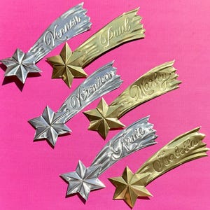 Könnte beinhalten: Eine Sammlung sternförmiger Ornamente mit Kometenschweifen, jeweils mit einem Namen versehen. Die Ornamente sind in Silber- und Goldtönen gehalten und vor einem rosa Hintergrund platziert. Namen sind 'Vernon', 'Paul', 'Veronica', 'Marley', 'Annie' und 'Isabella'.