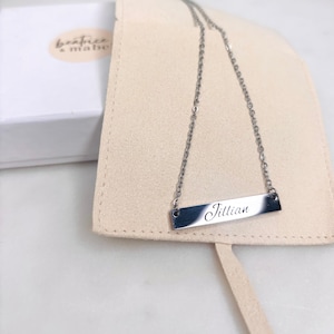 May include: A silver bar necklace with the name "Jillian" engraved on it. The necklace is displayed on a beige suede pouch, with a white box and the brand name "beatrice & mabel" in the background.