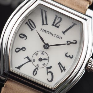 Puede incluir: Un reloj de pulsera Hamilton con caja plateada en forma de tonel y esfera blanca. La esfera presenta números arábigos negros, manecillas negras y una subesfera pequeña para los segundos en la posición de las seis. Incluye una correa de cuero.