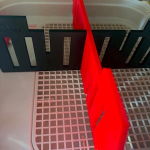 May include: A white plastic grid tray featuring two perpendicular dividers. One divider is black with rectangular cutouts and the word "series" embossed on the top left. The other divider is bright red and intersects the black piece at a right angle.