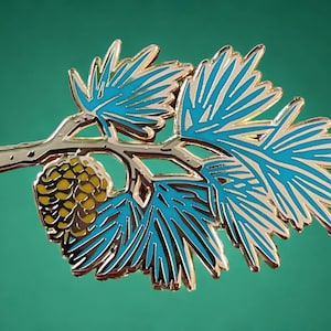May include: A gold-toned metal enamel pin shaped like a pine branch with bright turquoise needles and a textured yellow pinecone. The design features clean metallic outlines separating the colored enamel sections in a stylized botanical depiction.