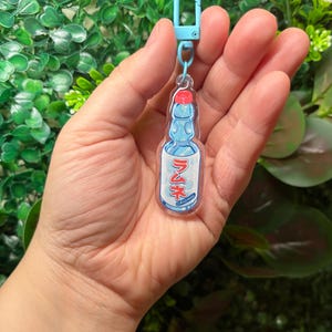 May include: A clear acrylic keychain shaped like a Ramune soda bottle, featuring a red cap and blue bottle design. The keychain has a light blue clasp. The bottle has Japanese text and the word "Original".