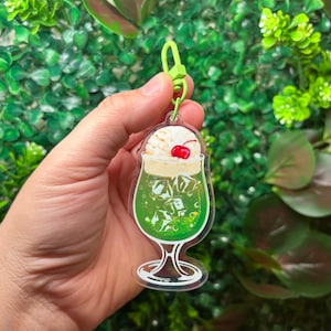 May include: A clear acrylic keychain featuring a green cocktail with ice cubes, whipped cream, and a cherry on top. The keychain has a green clasp and is held against a backdrop of green foliage.